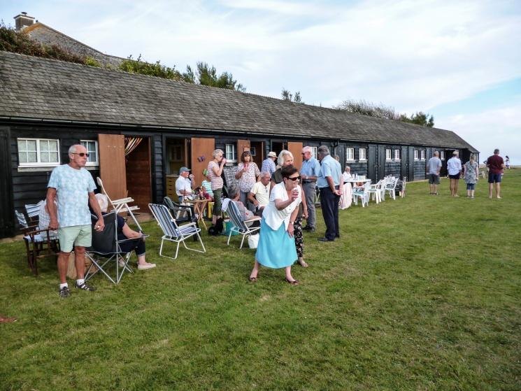 Twenty or more residents gathered on the grass in front of the long row of black timber beach huts, with folding chairs and tables out and the sea beyond