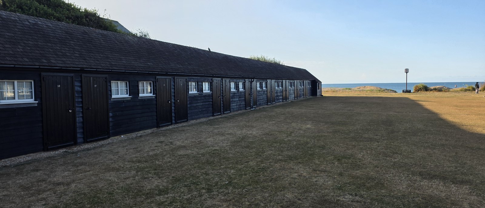 The long run of black timber beach huts along the edge of the mown Greensward, with the beacon and the sea to the right