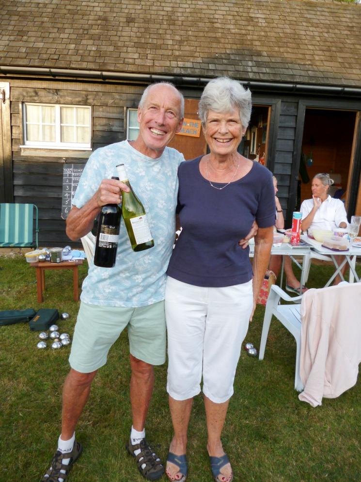 Two residents standing arm in arm in front of a beach hut, one holding two bottles of wine, with a chalked scoreboard on the hut wall behind them