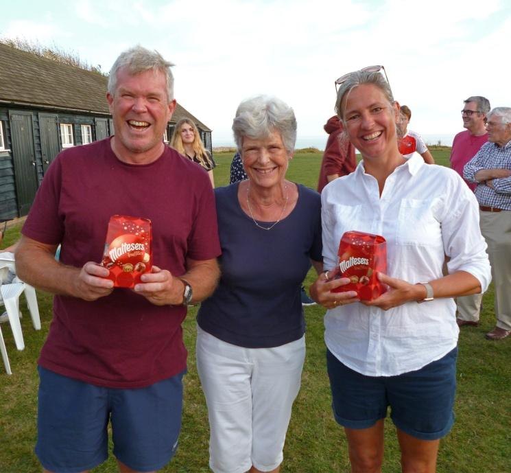 Three residents standing together on the Greensward holding boxes of chocolates, smiling at the camera, with the beach huts behind them
