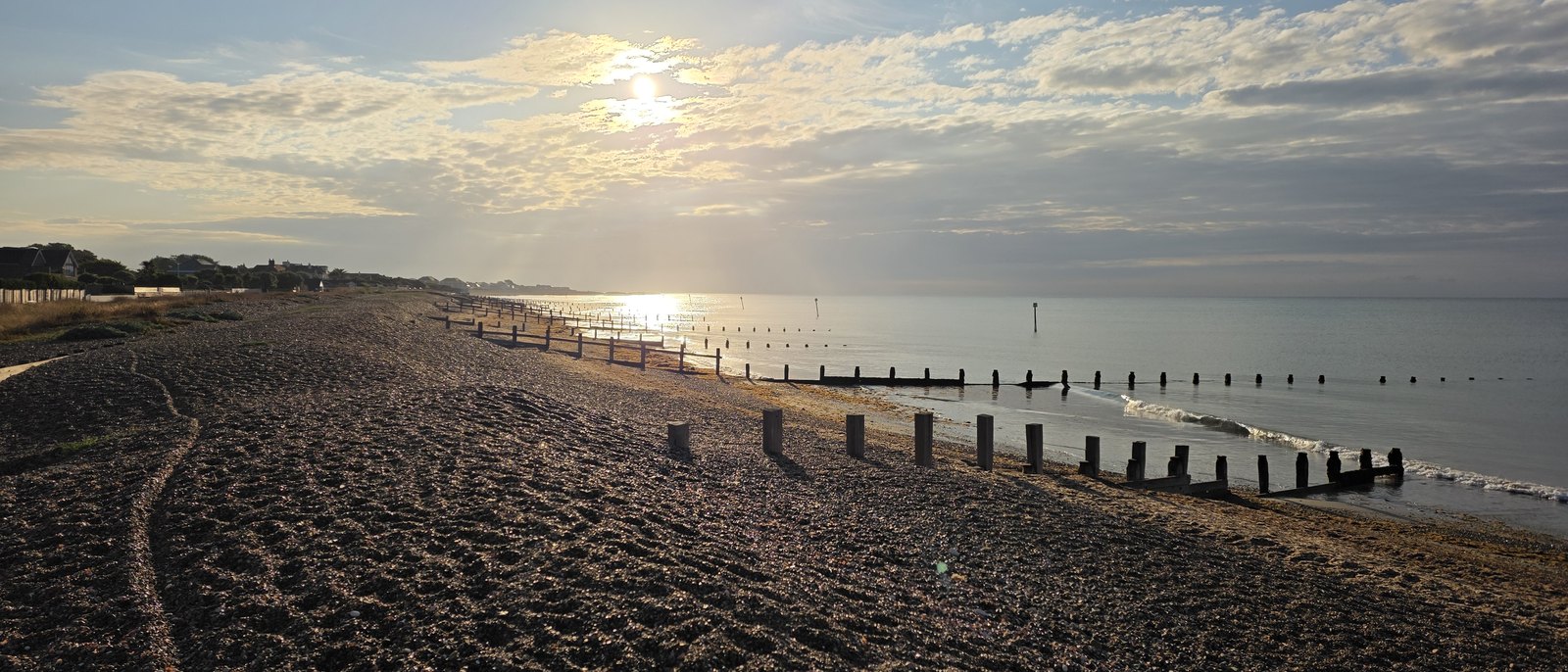 Early morning sun over a calm sea, with lines of timber groyne posts running out from the shingle beach and the rooftops of the estate on the skyline