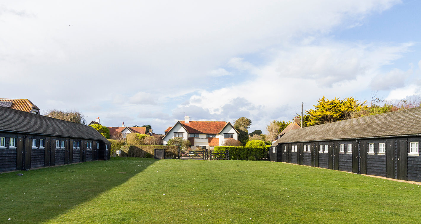A row of black timber beach huts along the edge of the Greensward, with mown grass in front of them and the sea beyond