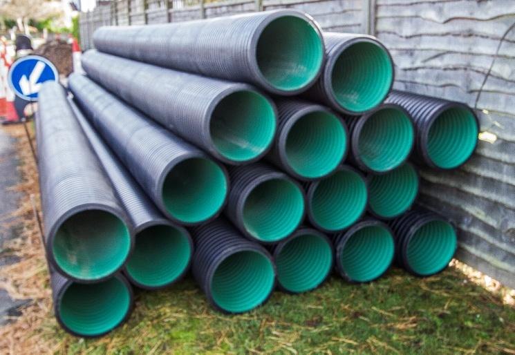 A stack of large black plastic land drainage pipes with green interiors, piled against a fence beside a road with a traffic sign