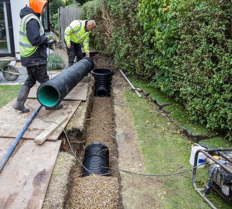 Two contractors in high visibility jackets laying a large corrugated drainage pipe into an open trench running down the side of a garden, with a petrol pump running beside them