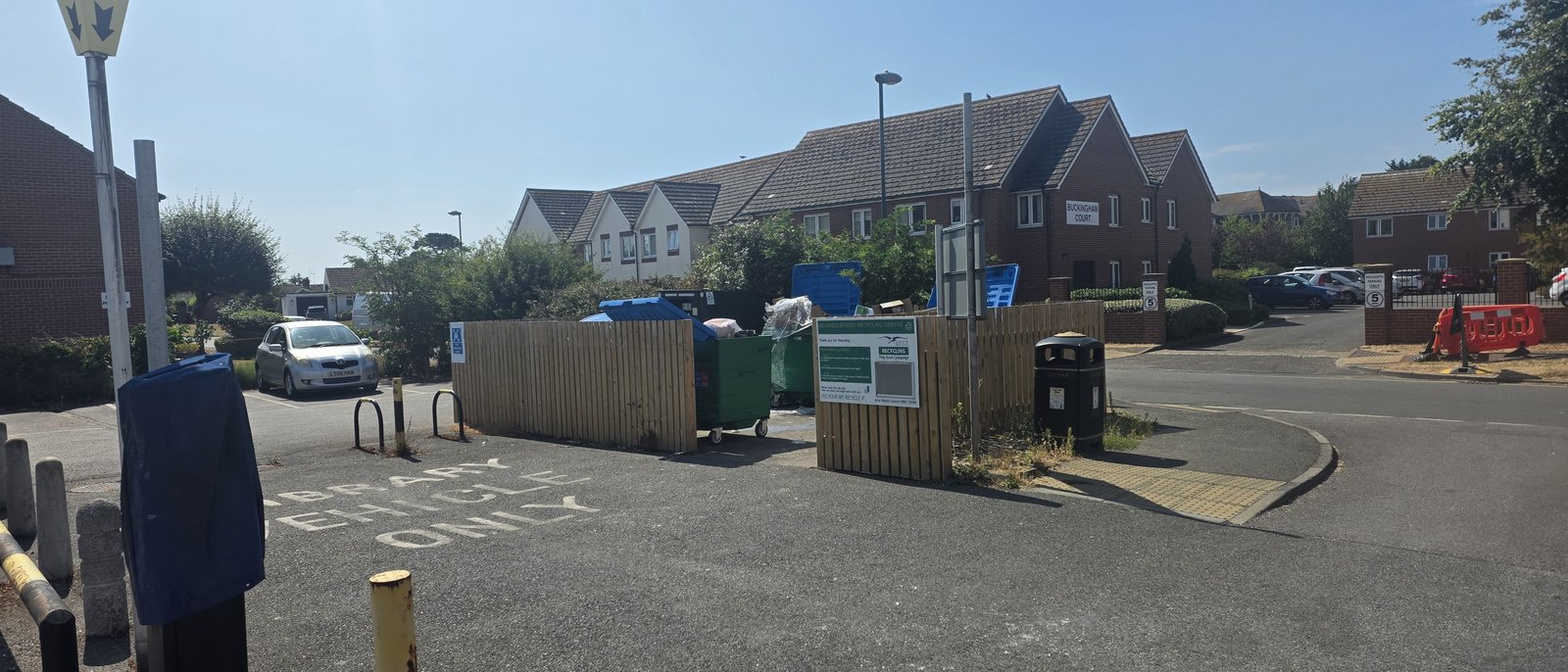 A fenced enclosure in a car park holding green and blue recycling bins, with an Arun District Council sign headed Neighbourhood Recycling Centre, and flats beyond