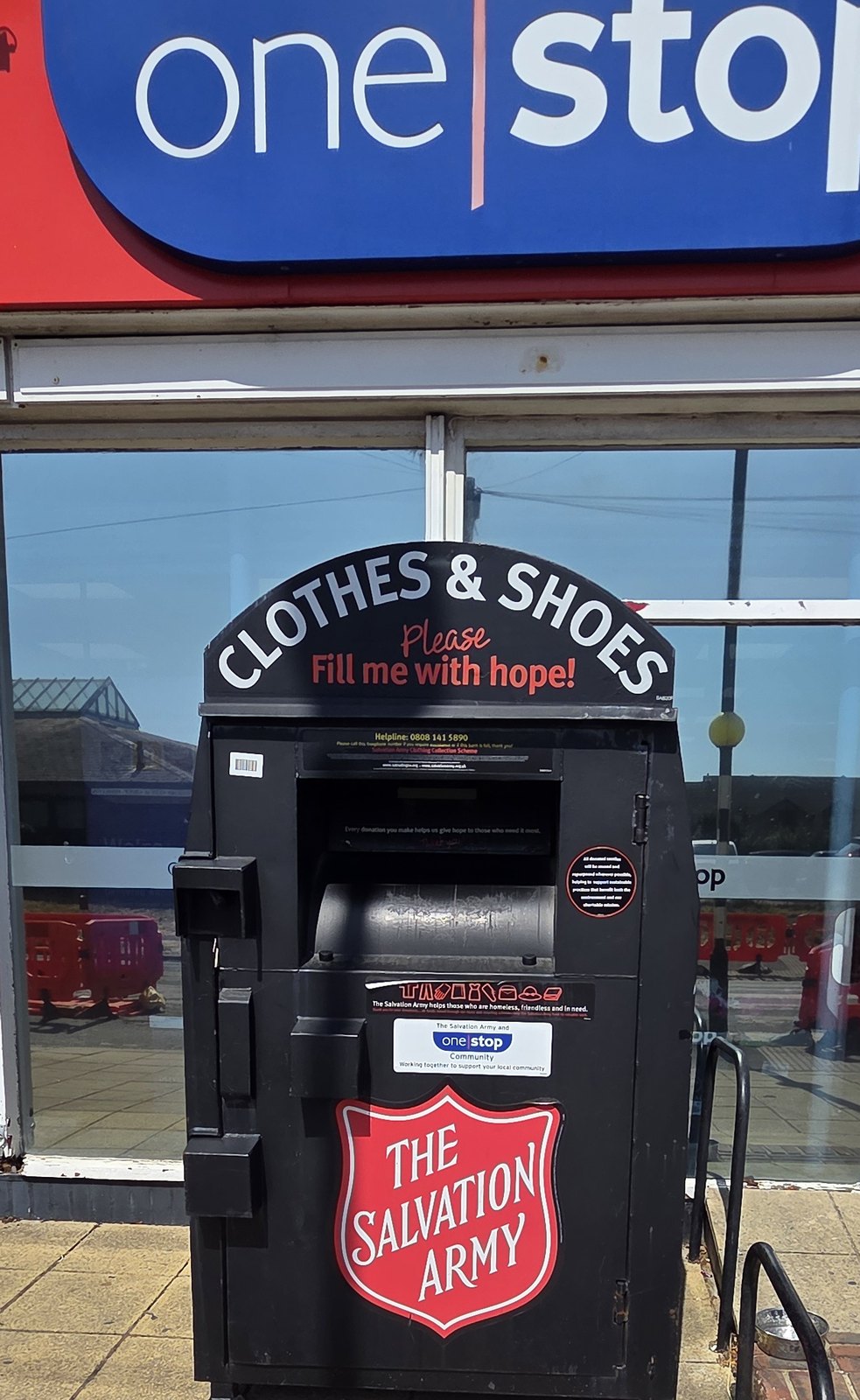 A black Salvation Army donation bank on the pavement outside the One Stop shop, with a curved top lettered clothes and shoes, please fill me with hope, and a Salvation Army shield on the front