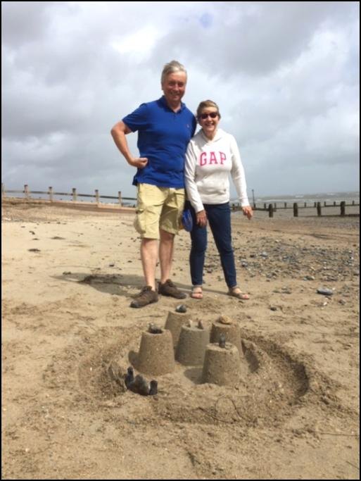 Two residents standing behind a completed sandcastle with four turrets on the beach, with groynes and a grey sea behind them