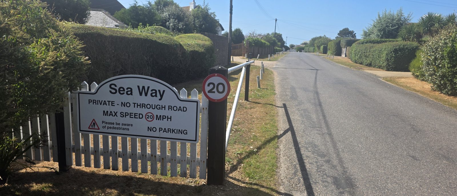 The entrance to Sea Way: a white sign on a picket fence reading Sea Way, private, no through road, max speed 20 mph, please be aware of pedestrians, no parking, beside a 20 mph roundel, with the road running away between grass verges and clipped hedges