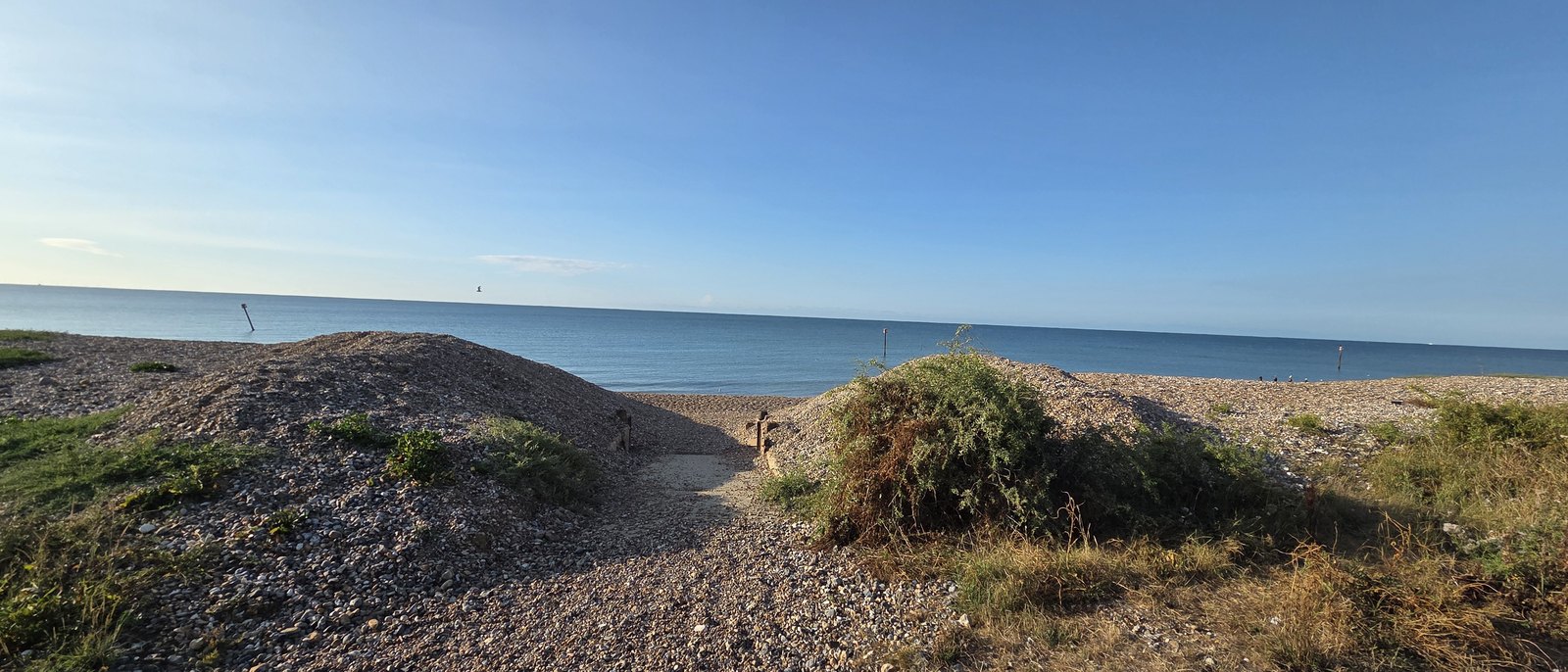 A track cut through the shingle bank down to the beach, with timber posts either side, the sea and marker posts beyond