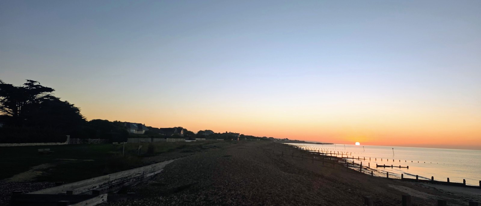 Sunrise over the Greensward and the sea, looking east along the shingle beach and its wooden groynes, with the cedar tree and rooftops in silhouette against the dawn sky