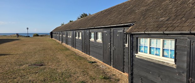 A terrace of black timber beach huts with numbered doors, the Greensward and the sea beyond