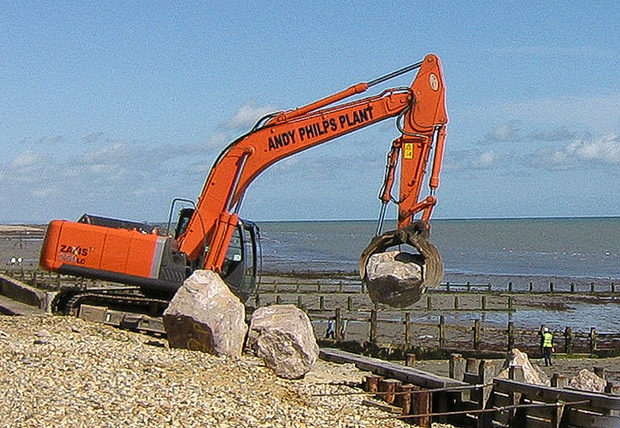 A tracked crane placing rock armour on the beach beside a timber groyne