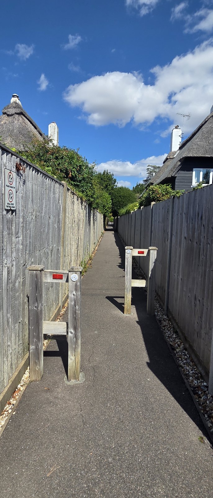 A narrow path between close-boarded fences with timber chicane barriers across it, a sign on the fence asking dog owners to clean up, and a thatched roof visible beyond