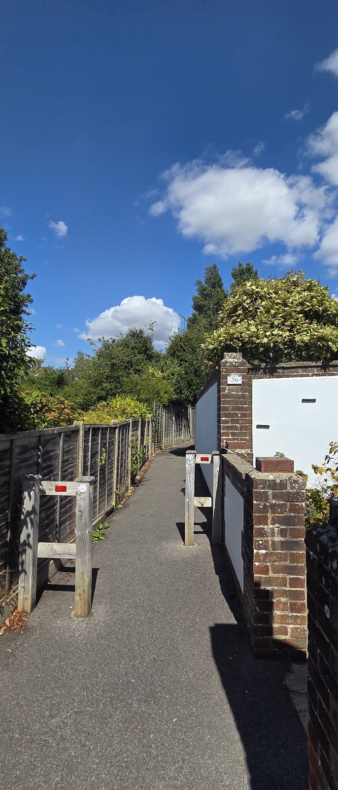 A narrow path with timber chicane barriers set across it carrying red reflective markers, a brick wall and a white rendered building beyond, under a blue sky with cumulus cloud